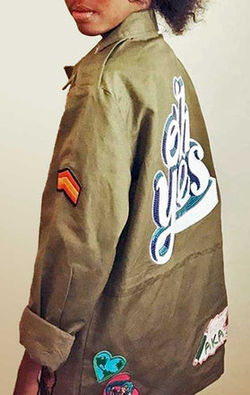 Load image into Gallery viewer, Child models back/side view of CdJ KidsRock jacket in solid army green with kid-friendly patch embellishments.
